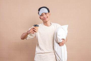 Young man in pajamas wear sleep mask, hold pillow and takeaway paper cup of coffee or tea isolated...