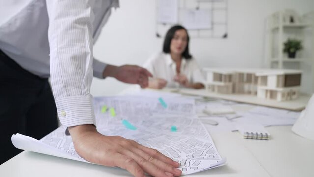 Skilled caucasian interior designer working with house model while discussing with asian architect engineer about house plan on meeting table with blueprint and house model scatter around. Immaculate.
