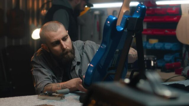 Male craftsman pulls strings on guitar and sets up their sound. Woodworking master makes wooden musical instrument in modern workshop. Colleague works in the background. Handmade and entrepreneurship.