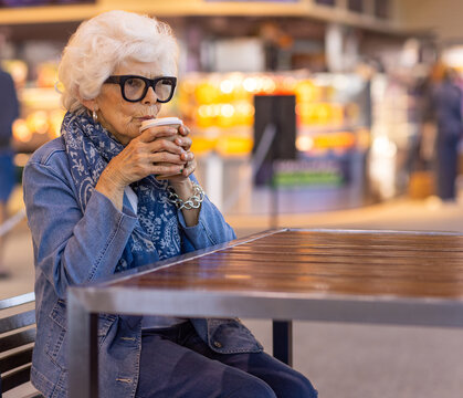 white-haired lady wearing denim sitting at table sipping coffee from takeaway cup