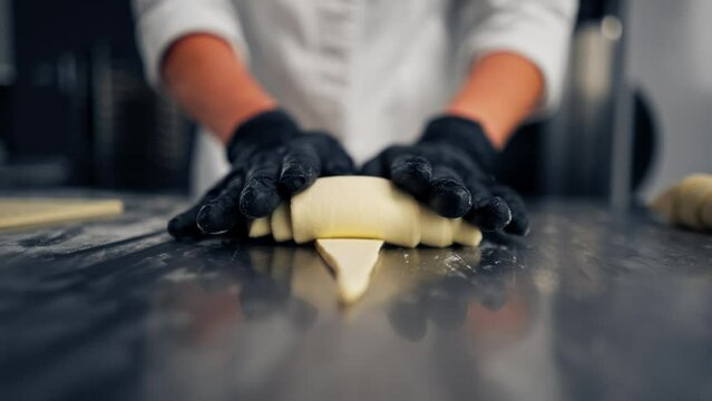 Close-up of the chef's gloved hands shaping and twisting raw dough into shape of croissants for baking