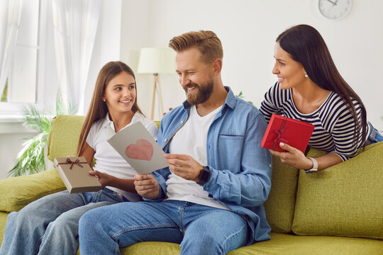 Cheerful father sitting on sofa at home with hir girl child and wife and reading handmade greeting card with heart during holiday celebration fathers day. Happy man dad receiving present from family.