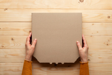 Obraz premium Pizza closed carton box in human caucasian hands on natural wooden table flat lay mockup with blank space