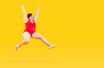 Fototapeta na wymiar Cheerful fat woman in swimsuit rejoices, jumps and has fun while enjoying summer vacation. Funny chubby woman in red swimsuit hangs in air near copy space on yellow background. Banner. Full length.