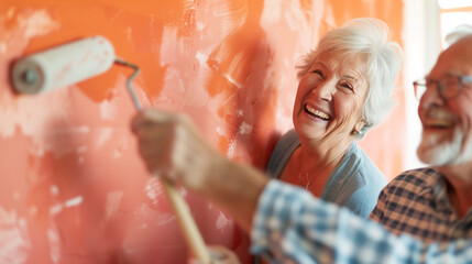 A senior woman with white hair laughs joyfully while painting a wall orange, as a senior man with glasses shares the moment with her, both showing the happiness of working together