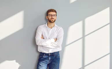 Confident bearded man in eyeglasses posing with crossed arms at gray wall. Portrait of handsome...