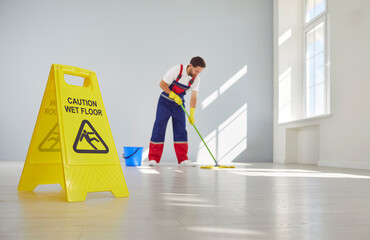 Caution wet floor sign with male young cleaner mopping empty room or office in background. Man...
