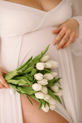 Obraz na płótnie Canvas regnant woman in a beautiful white dress holding her belly with one hand a bouquet of yellow tulips in the other hand. Close up photo of pregnant belly