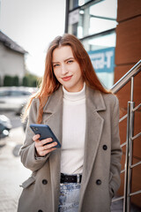 
a beautiful girl in a white turtleneck and a gray coat with red hair holds a phone in her hands