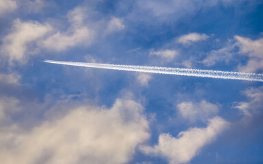 White trail of plane flying among white clouds in blue sky