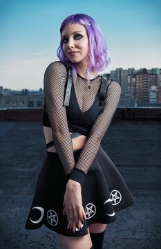 Portrait of cute smiling gothic girl on roof at evening time. Pastel goth with violet (purple) hair in black clothes