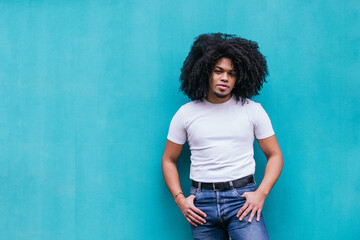 Casual yet confident Afro-Latino man with curly hair standing against a light turquoise wall,...