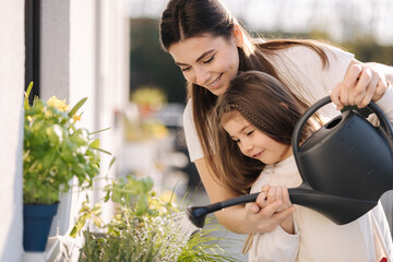 Happy daughter watering plants with her beautiful mom. Girl's in bright overall gardening on balcony