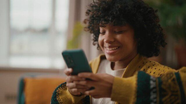 Young woman sitting with phone. African American woman hold in hand use mobile phone typing messages. Girl joy spend free spare time in living room. Communication on social networks, rest concept.