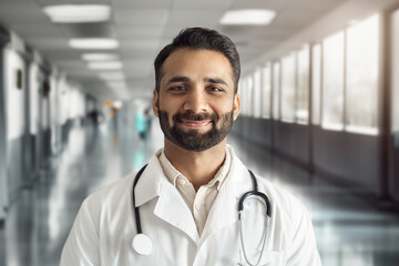 Headshot portrait of smiling mature indian doctor wearing white coat, stethoscope looking at camera standing in hospital corridor. Therapist or general practitioner, medical staff, clinic presentation
