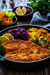 Crispy breaded fried cutlet with baked potatoes and cooked red cabbage on wooden table
