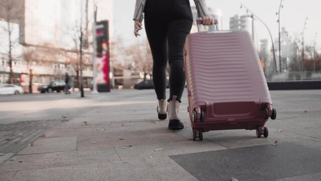 Woman walking on a city street and rolling a wheeled suitcase behind her close up, view from behind. Concept of travel, tourism and enjoying a trip