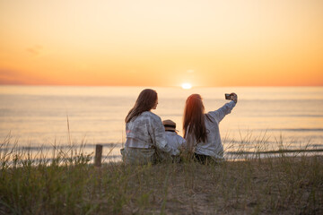 Back view of two caucasian female friends and a child making selfie at sunset on the Baltic seashore
