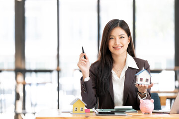 Real estate agent with model of house sitting at office, smiling to camera.