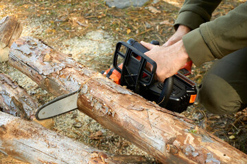 Male hands seen holding an electric chainsaw cutting through a thick log of wood  - obrazy, fototapety, plakaty