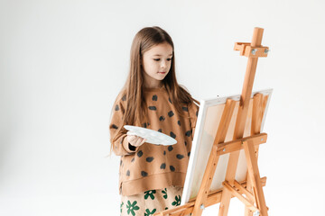 a 6-year-old girl with a brush in her hands in front of an easel with a canvas on an isolated white...