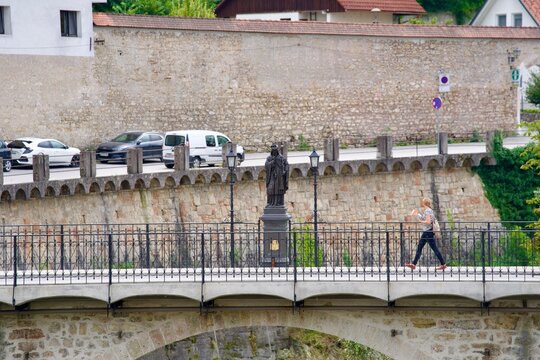 Woman crossing medieval bridge over Selca Sora River with sculpture of Saint Nepomuk at City of Skofja Loka on a cloudy summer day. Photo taken August 9th, 2023, Skofja Loka, Slovenia.