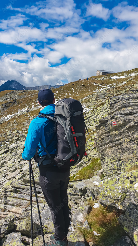 Poster Hiker man on alpine terrain with panoramic view of remote cottage Hagener Huette in High Tauern National Park, Carinthia, Austria. Idyllic hiking trail in Austrian Alps. Wanderlust paradise Mallnitz