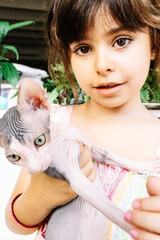 6-year-old Spanish girl looking at the camera while holding a purebred Sphinx cat in her arms. Vertical.