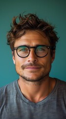 Obraz premium A man with tousled hair, wearing glasses, looks forward, against a teal background, appears contemplative