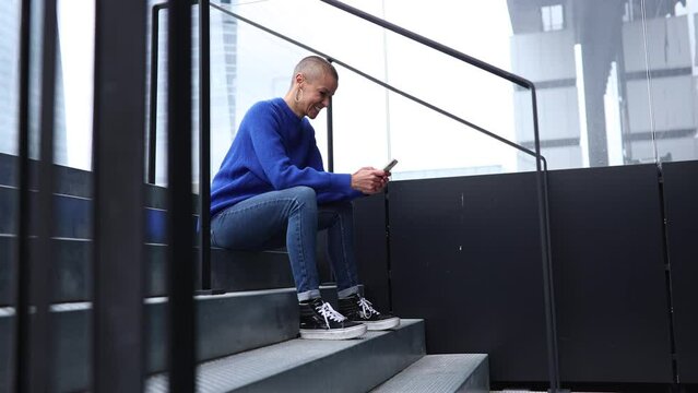 modern shaved bald woman sitting on stairs and gossiping online while laughing and texting with skyscrapers in the background