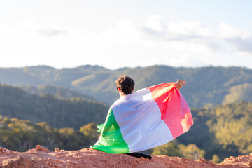 Italian man seated atop a mountain, proudly holding the Italian flag, surrounded by a mountainous landscape. Celebrating national pride during sports events or in commemoration of Italy's National Day