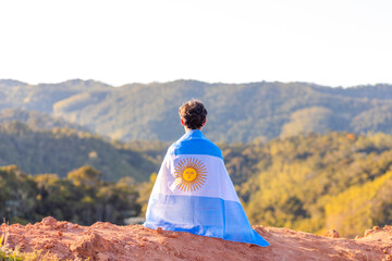 Argentinian man seated atop a mountain, proudly holding the Argentine flag, surrounded by a...