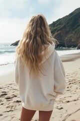 Fototapeta na wymiar Junge Frau mit blonden Haaren in einem weißen Kapuzenpullover mit dem Rücken zur Kamera am Strand