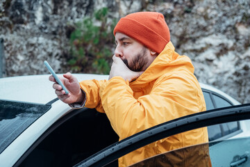 Young bearded man using phone while resting stop on road trip. He is wearing casual yellow jacket and red hat. Travel by car.