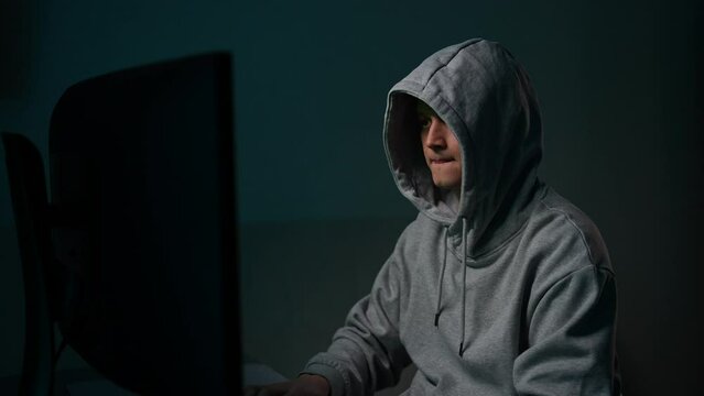 Hacker man in gray hoodie typing on keyboard, preparing to break password, drinking coffee. Programmer male writing code, virus program for cyber attack using computer, drinking beverage in dark room.