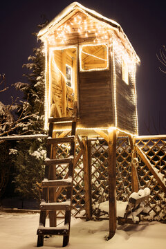 A wooden treehouse on a winter night - covered in snow and lit up with lots of Christmas lights.