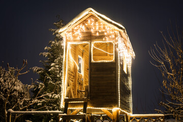 A wooden treehouse on a winter night - covered in snow and lit up with lots of Christmas lights.