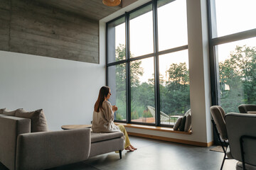 Woman in a country house or hotel with panoramic windows in pine forest sitting on the bed and enjoys beautiful view with light sun. Good morning and recreation on nature concept