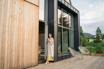 Young woman staying on terrace of wooden modern house with panoramic windows near pine forest....