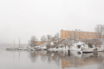 Scandinavian style historic yellow stone buildings with white snow on the ground on Skeppsholmen, Stockholm, Sweden by the water with a small boat marina during heavy snowfall in winter