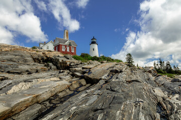 Pemaquid Point Lighthouse and Bell Tower in Bristol Maine sits atop rugged rock layers on a bright summer day dotted with puffy clouds