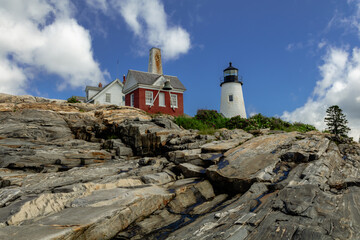 Pemaquid Point Lighthouse and Bell Tower in Bristol Maine sits atop rugged rock layers on a bright summer day dotted with puffy clouds