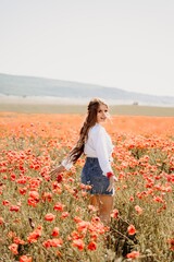 Obraz na płótnie Canvas Happy woman in a poppy field in a white shirt and denim skirt with a wreath of poppies on her head posing and enjoying the poppy field.