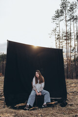 A beautiful sad girl sits on a chair, on a black background, set among deforestation at sunset. She closed her eyes. The wind blows your hair