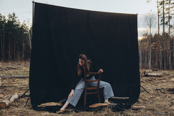 Girl, woman with bare feet sits on a chair on a black background set among deforestation. All...
