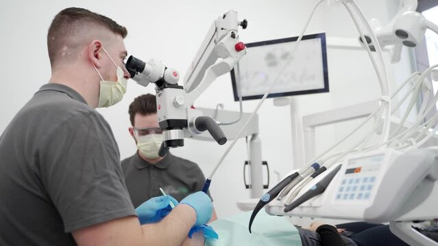 Male dentist with assistant operating patient tooth using microscope at modern dental clinic. Oral surgeon cleans root canal and cauterizes the dental nerve. Endodontist treat patient's root canals.