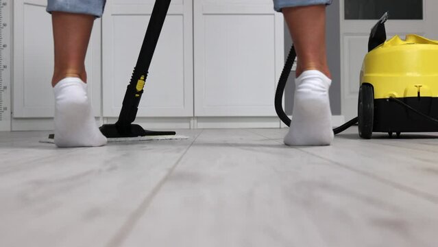 girl in white socks steps back, washes light floor in front of her with professional steam vacuum cleaner. Cleanliness and disinfection in house with the help of the services of a cleaning company