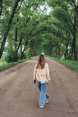 A stylish girl, a woman walks through a tunnel of green trees. Warm summer day. The girl, lightly dressed, walks off into the distance. Clothes flutter in the wind