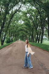 A young stylish girl, a woman, walks through a tunnel of green trees. Warm summer day. The girl is dressed lightly. Clothes flutter in the wind