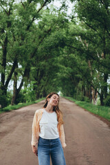 A stylish girl, a woman stands in a tunnel of green trees. Warm summer day. The wind blows the girl's hair and clothes. The girl is happy